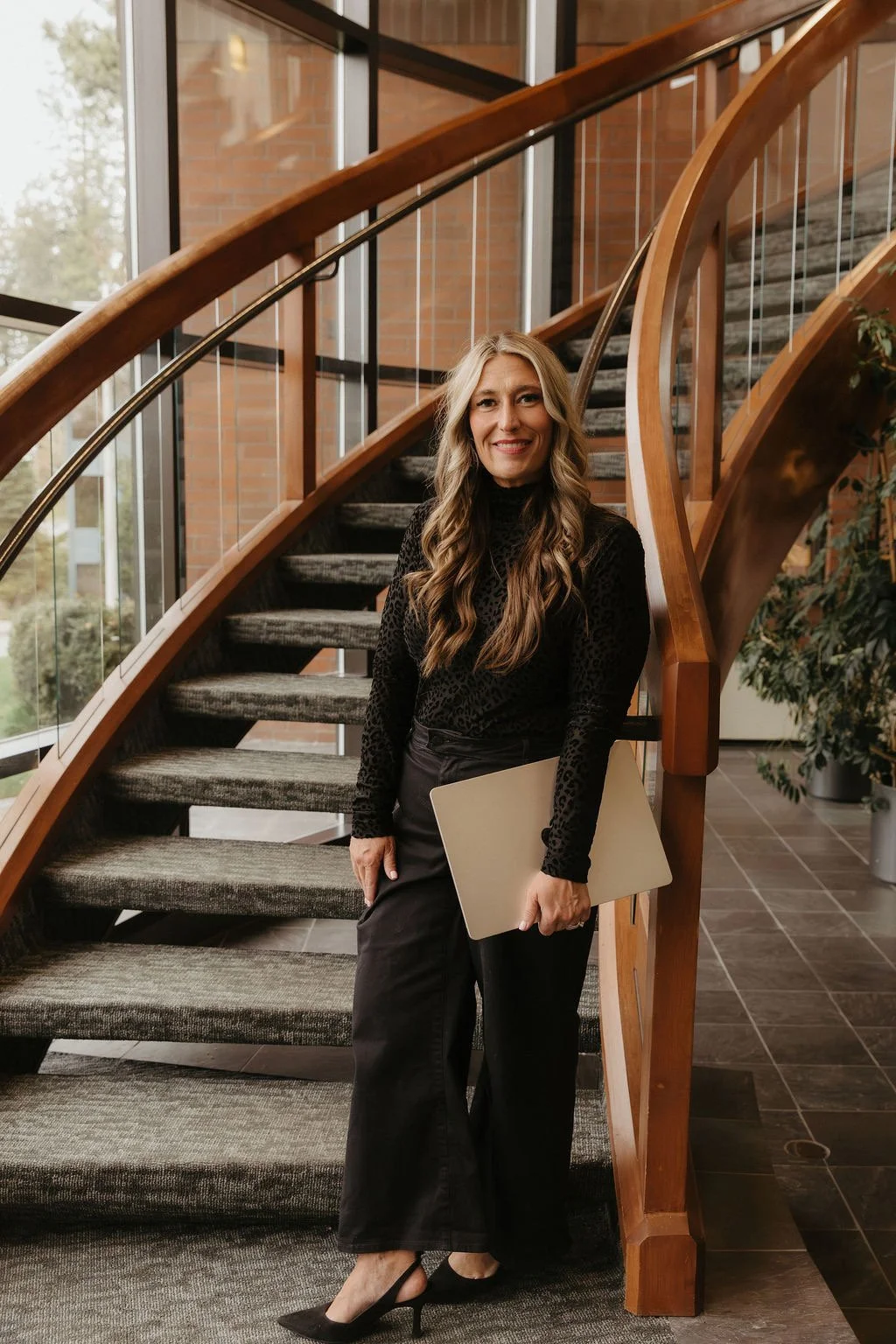 A woman with long wavy blonde hair, wearing a black patterned top and black pants, holding a laptop, standing on a carpeted staircase inside a modern building with large windows and brick walls.