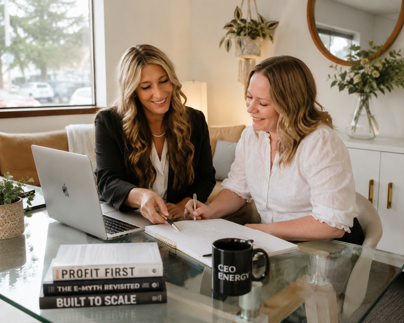 Two women sitting at a glass table, reviewing documents together with a laptop, a book titled 'Profit First', and a coffee mug that says 'CEO Energy'. They are smiling and engaging in a discussion in a well-lit room with plants and a large window.