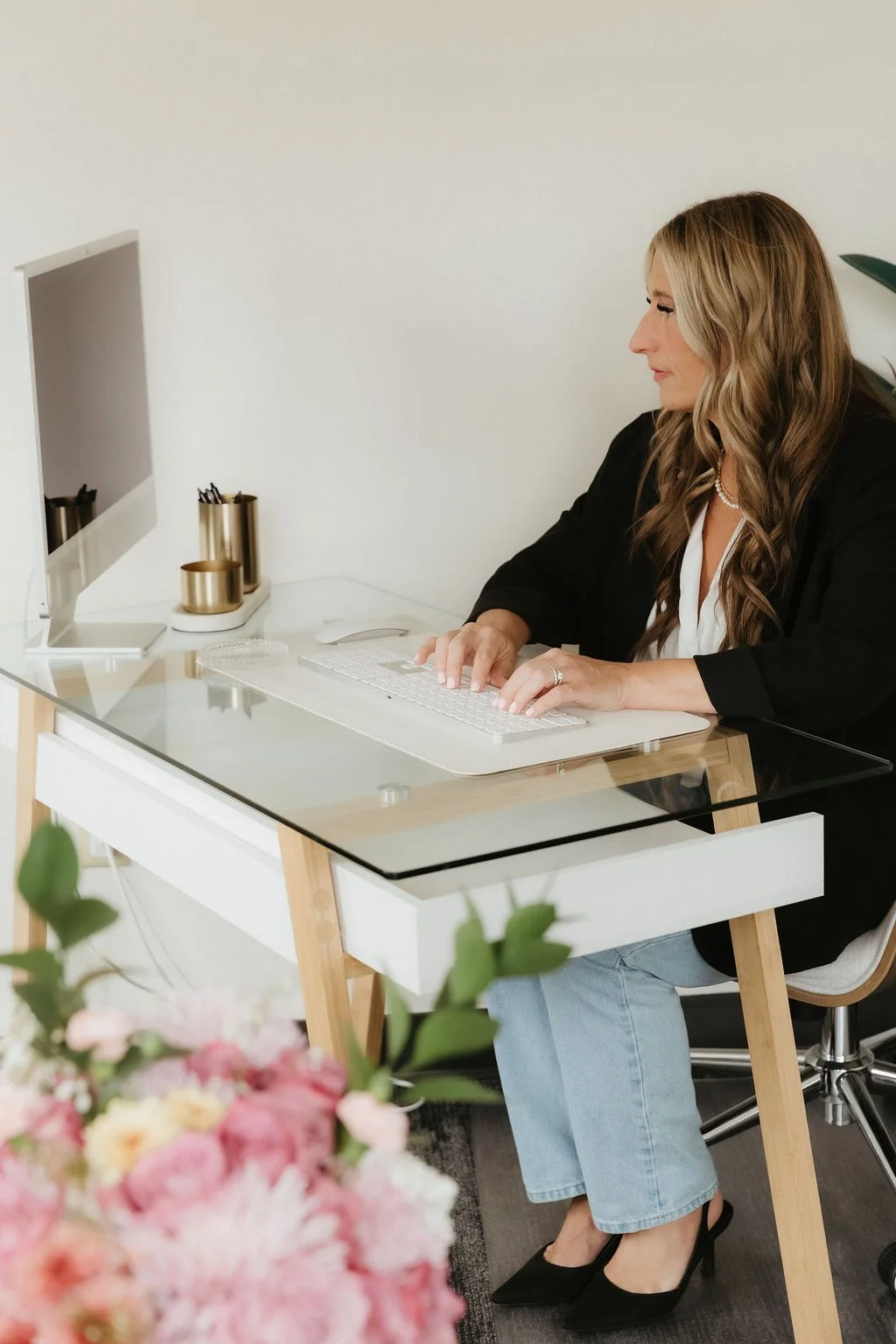 Woman working at a glass desk with a computer, flowers in the foreground, minimal office decor.