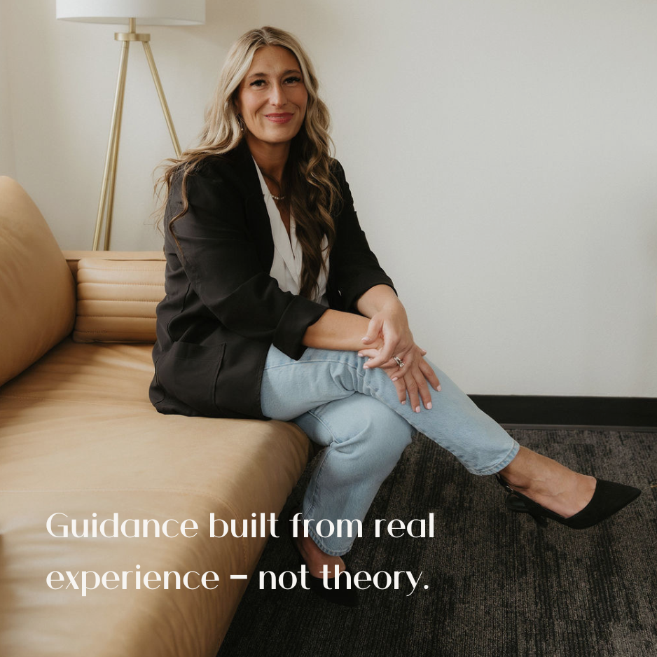 A woman with long wavy blonde hair is sitting on a beige sofa in a modern room. She is wearing a black blazer, white top, light blue jeans, and black heels, smiling at the camera. There is a floor lamp behind her and plain white walls.