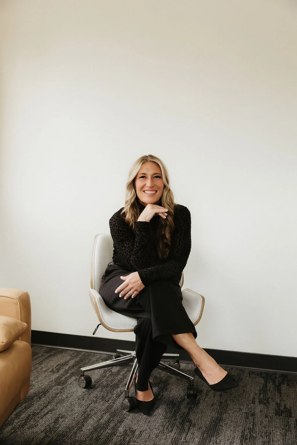 A woman with long blonde hair sitting on a white office chair, smiling with her hand resting under her chin, wearing a black long sleeve top and black pants, in a room with beige carpet, white wall, and a beige couch.