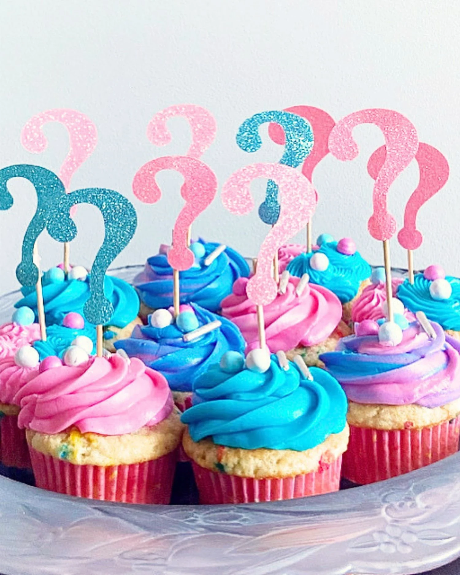 Decorated Gender Reveal cupcakes with pink and blue frosting, topped with sparkly number and question mark toppers, arranged on a silver tray.