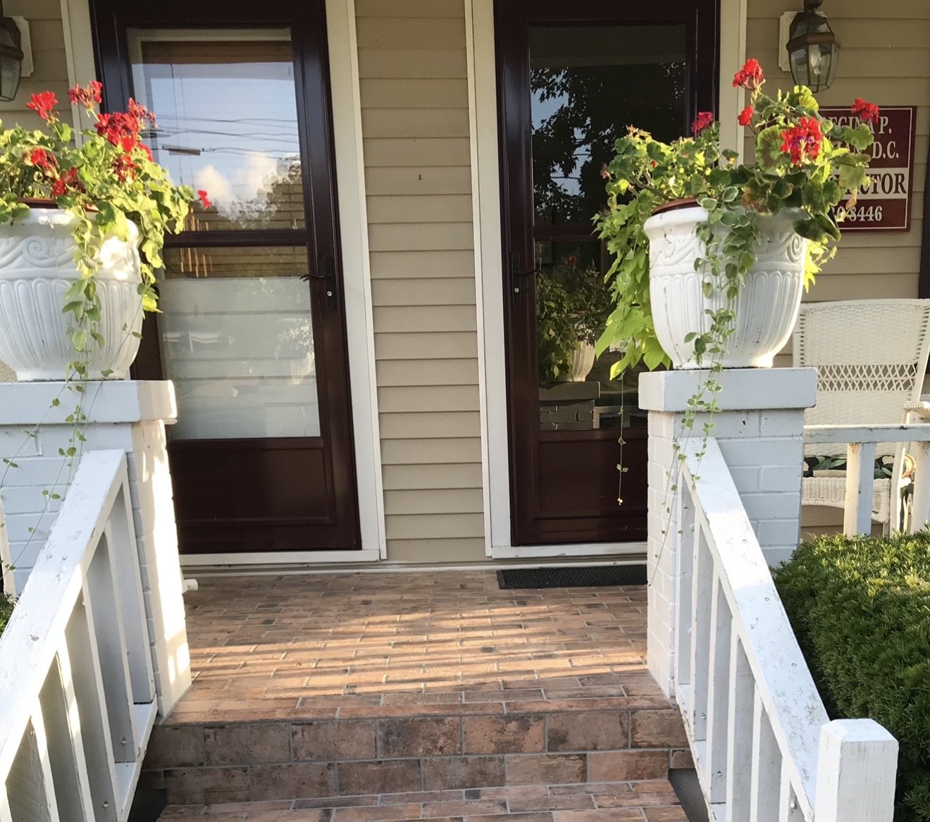 Front porch of a house with brick steps, white railing, two large flower pots with red and pink flowers, and a door with a screen