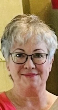 A middle-aged woman with short, curly gray hair and glasses, smiling at the camera.