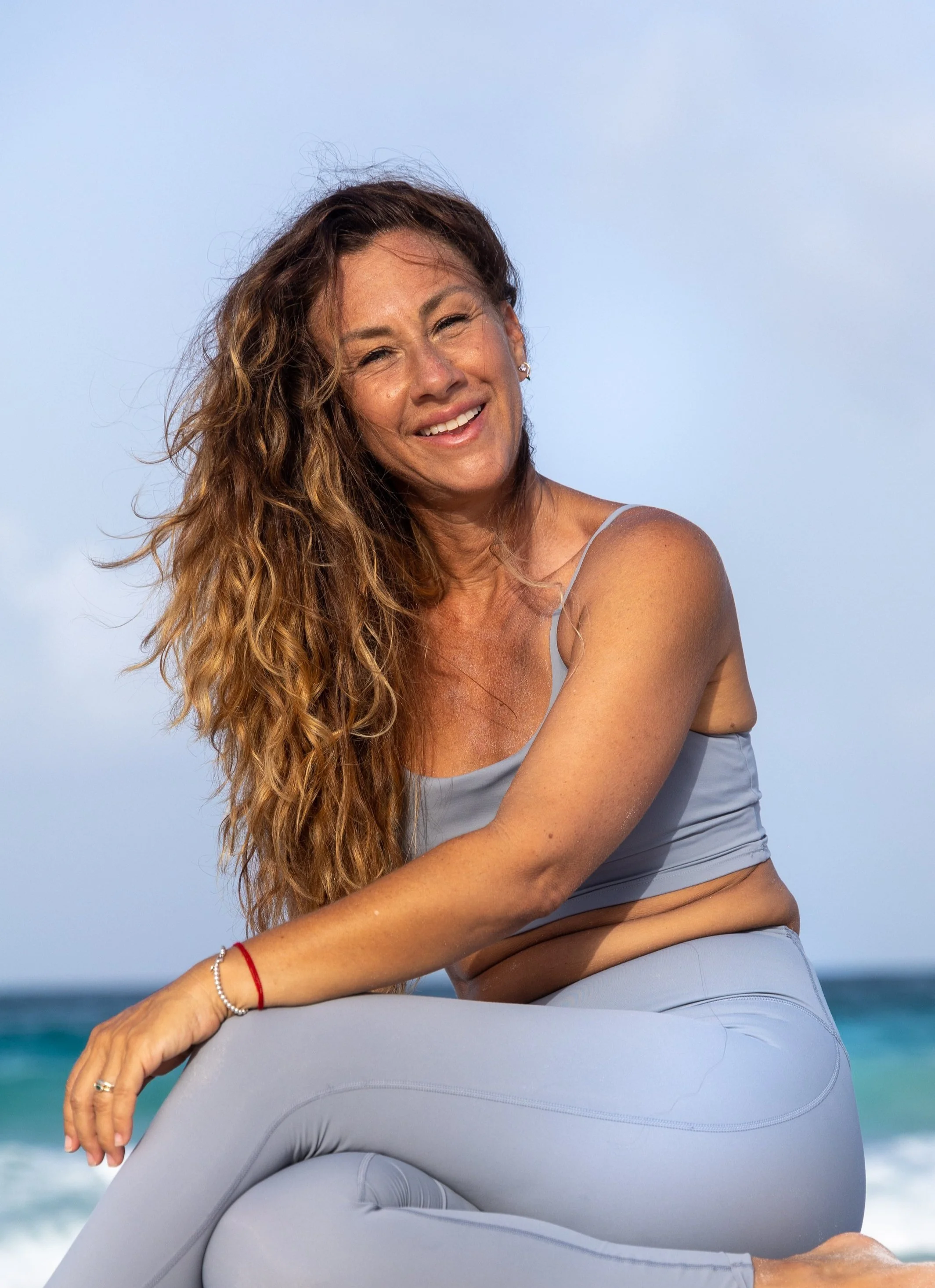 A woman with long curly hair smiling at the camera, wearing a light blue sports bra and leggings, sitting on a large inflatable object near the beach with ocean in the background.