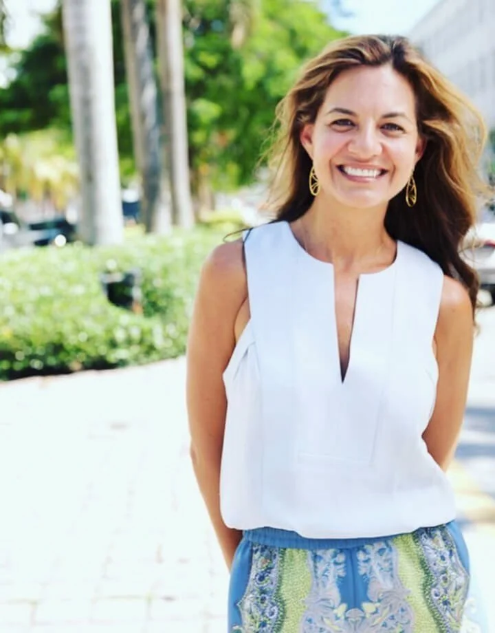 A woman smiling outdoors on a sunny day, wearing a white sleeveless top and patterned blue and green skirt, with earrings and brown hair.