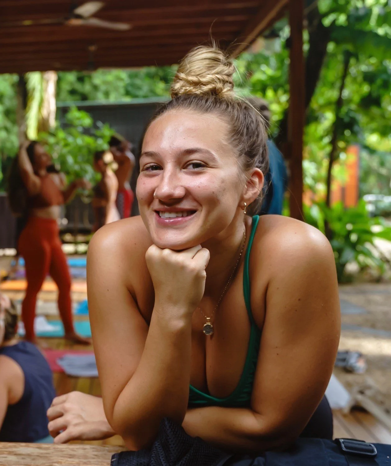 A smiling woman with blonde hair tied in a messy bun, wearing a green tank top, resting her chin on her hand, in an outdoor setting with lush green plants and other people in the background.