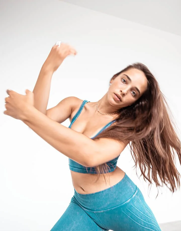 Young woman with long brown hair wearing blue sports bra and leggings, stretching or dancing against a plain white background.