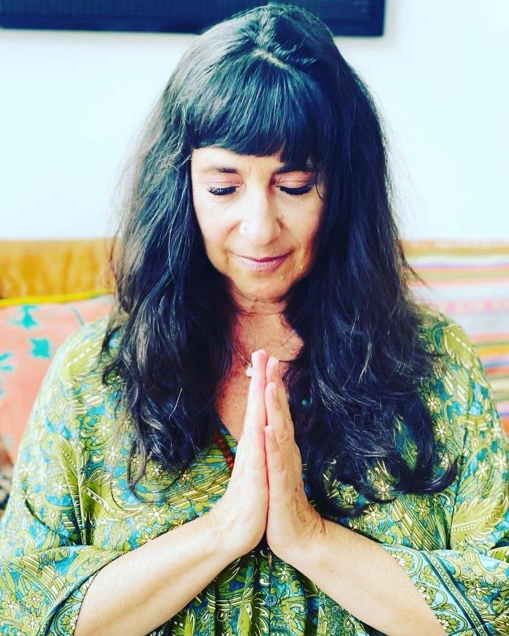 A woman with long dark hair and bangs eyes closed, hands pressed together in a prayer pose, wearing a colorful, patterned top indoors.