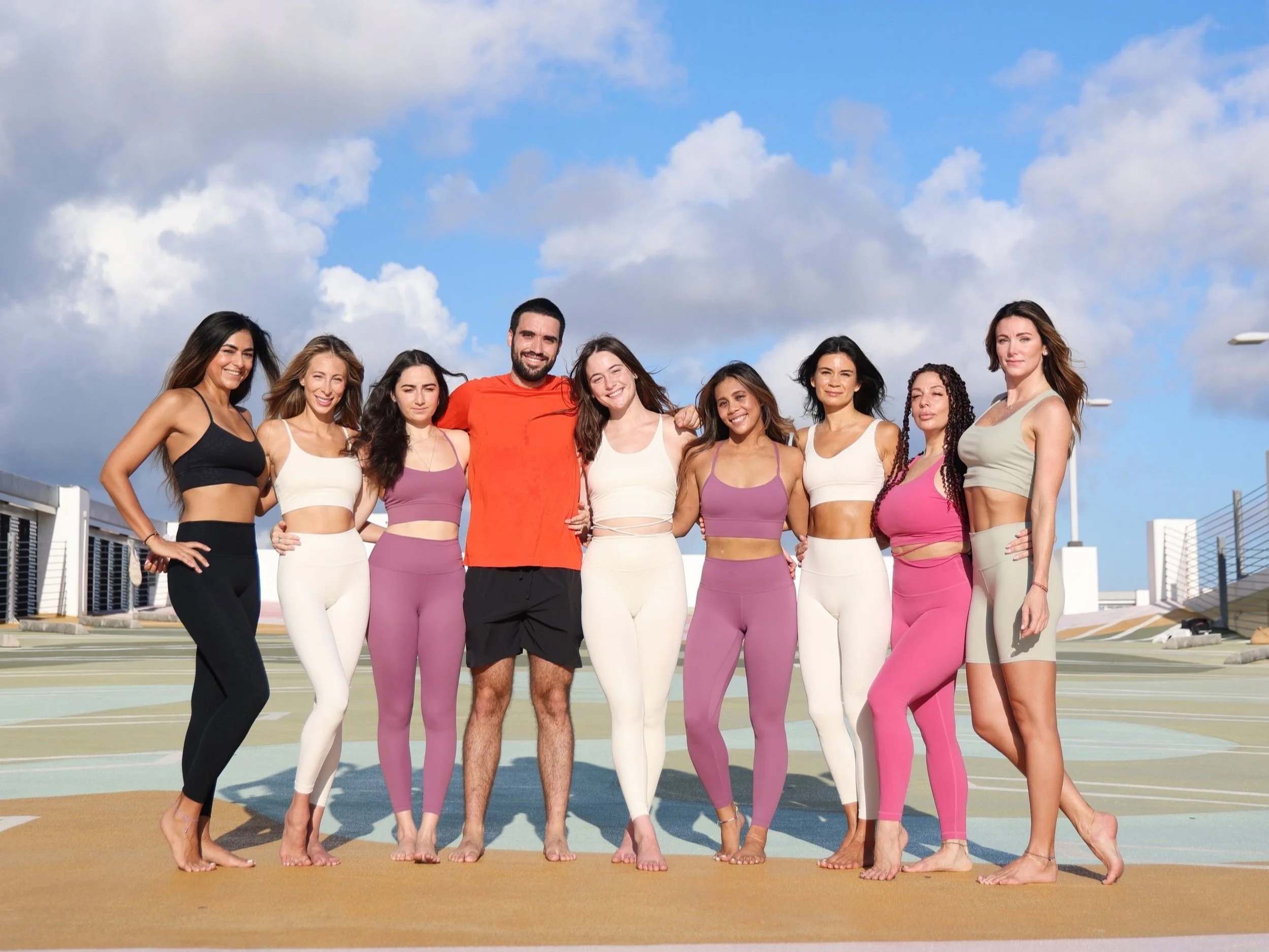 A group of ten young adults, nine women and one man, standing together on a rooftop race track during the daytime. The women are dressed in colorful athletic wear, while the man is wearing a red shirt and black shorts. They are smiling, with their arms around each other, under a partly cloudy sky.