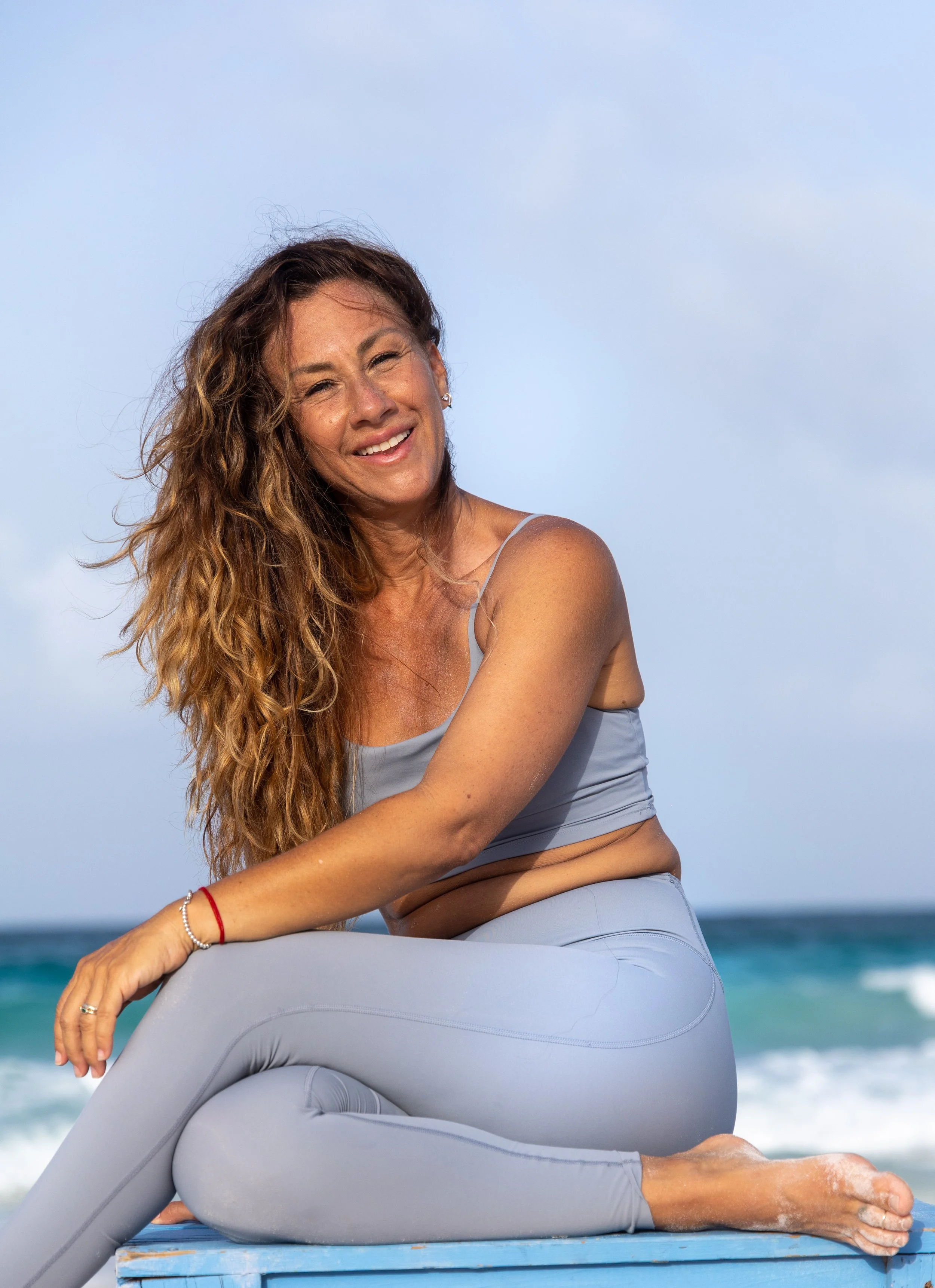 A woman with long curly hair doing yoga on a yoga mat outdoors on the beach, with the ocean and sky in the background.