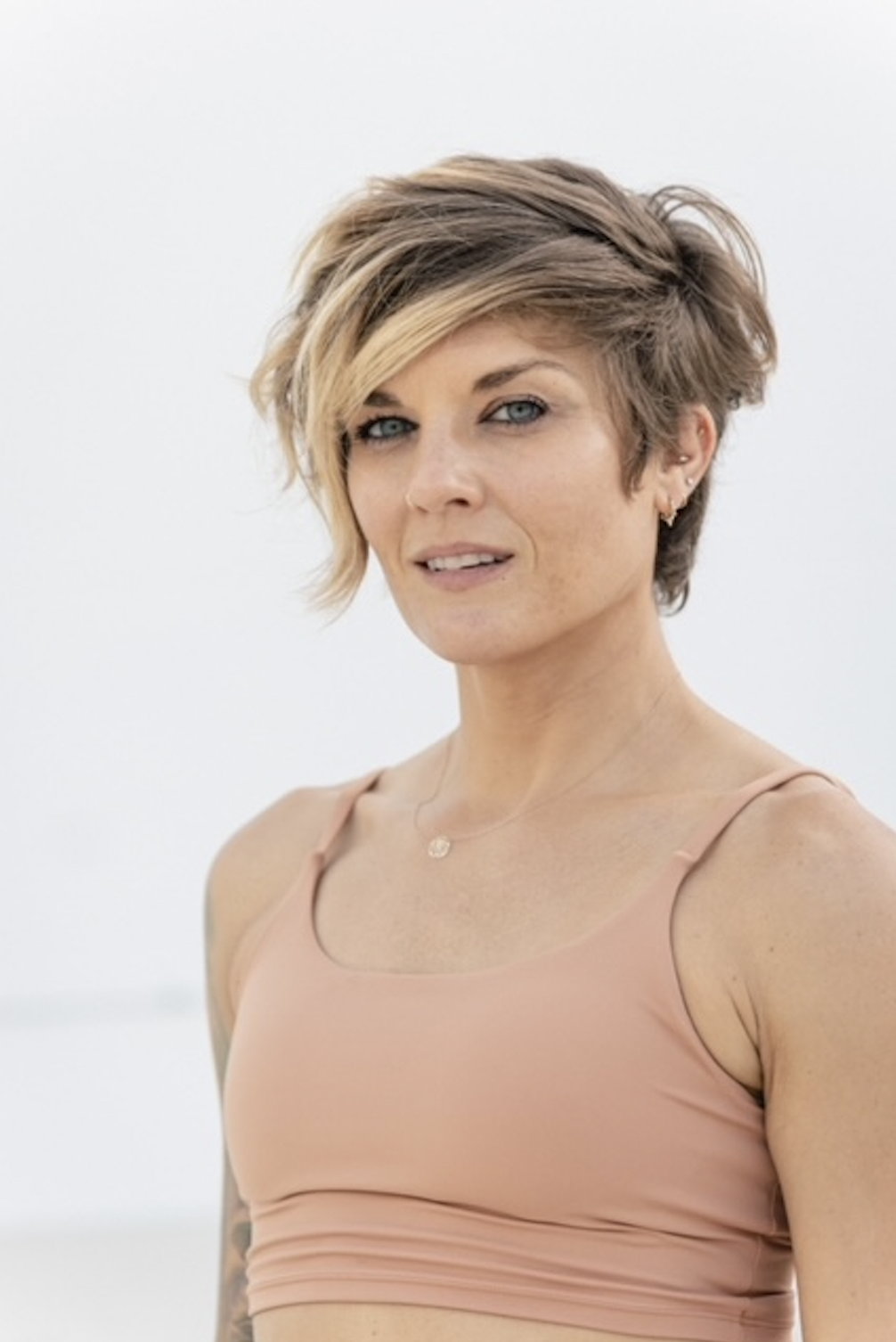 A woman with short, styled blonde hair and blue eyes wearing a light pink sports bra and a delicate necklace, standing against a plain light background.