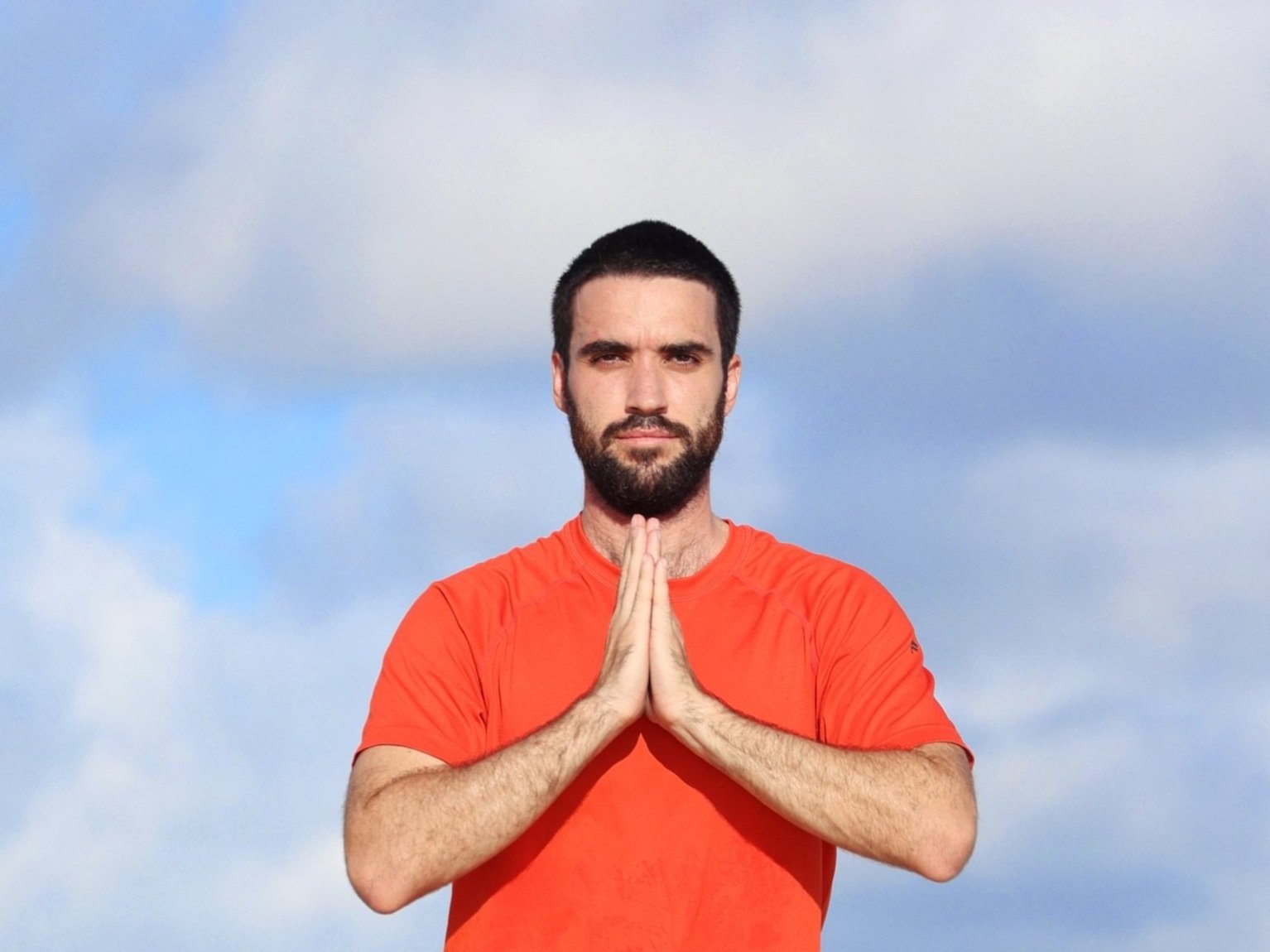 A man with dark hair and a beard, wearing an orange shirt, standing against a cloudy sky. He has his hands pressed together in front of his chest in a prayer or meditative pose.
