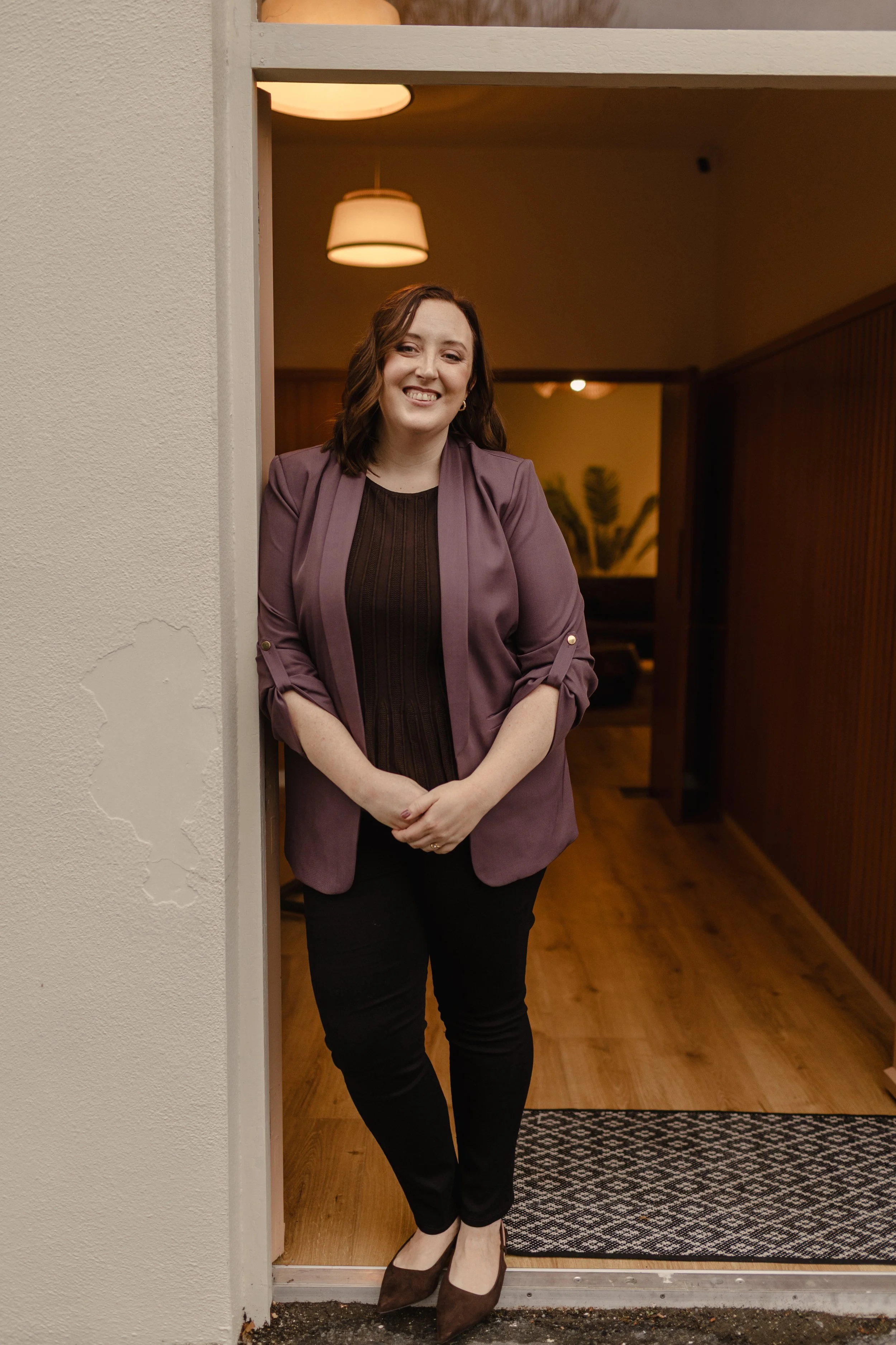 A woman with shoulder-length brown hair, wearing a mauve blazer over a dark top, black pants, and brown heels, stands in a doorway smiling at the camera inside a warmly lit room.
