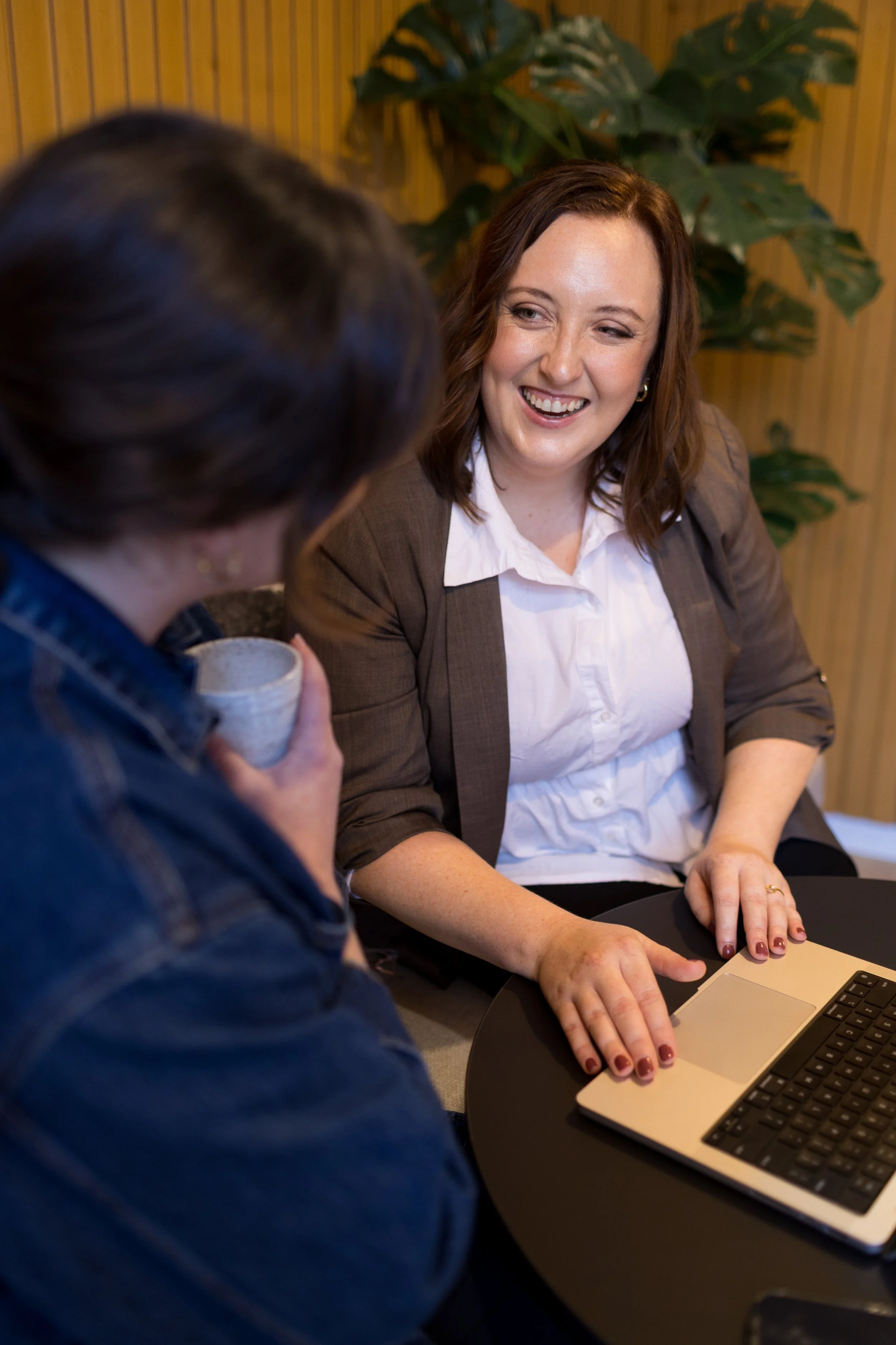 A woman with brown wavy hair, dressed in a white shirt and a brown blazer, smiling and talking to a person with dark hair, who is holding a coffee cup. They are seated at a black table with a laptop, in a room with wooden walls and a large green plant in the background.