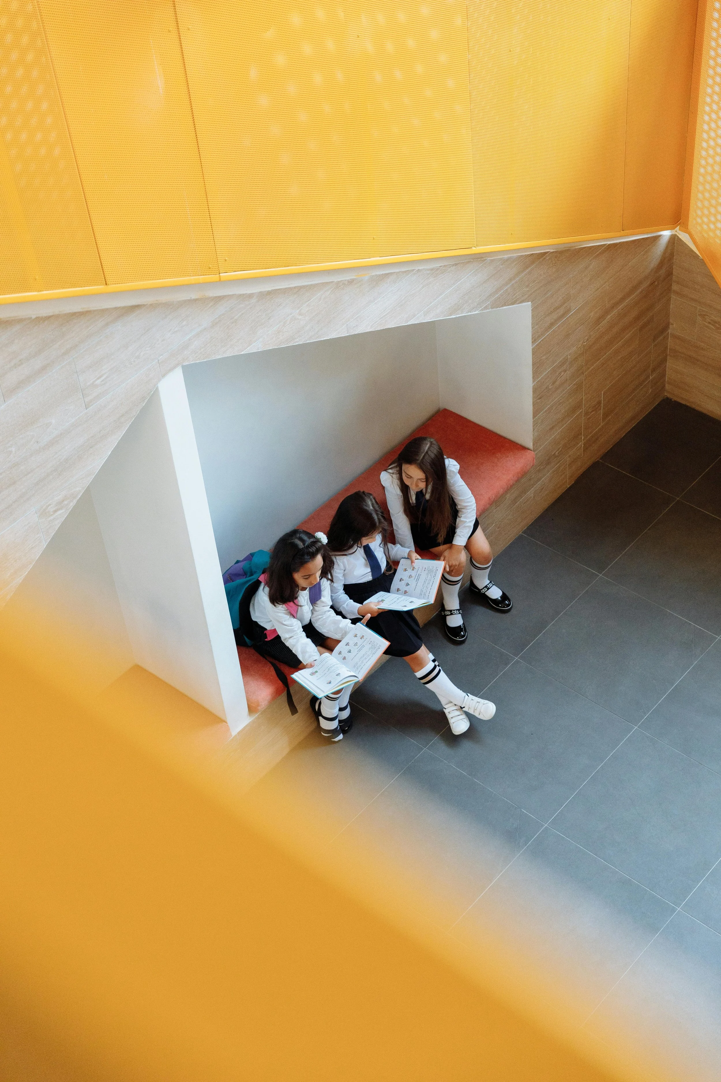 Four students sitting on a built-in bench against a wall, looking at a book together, in a school corridor with yellow and wood-paneled walls.