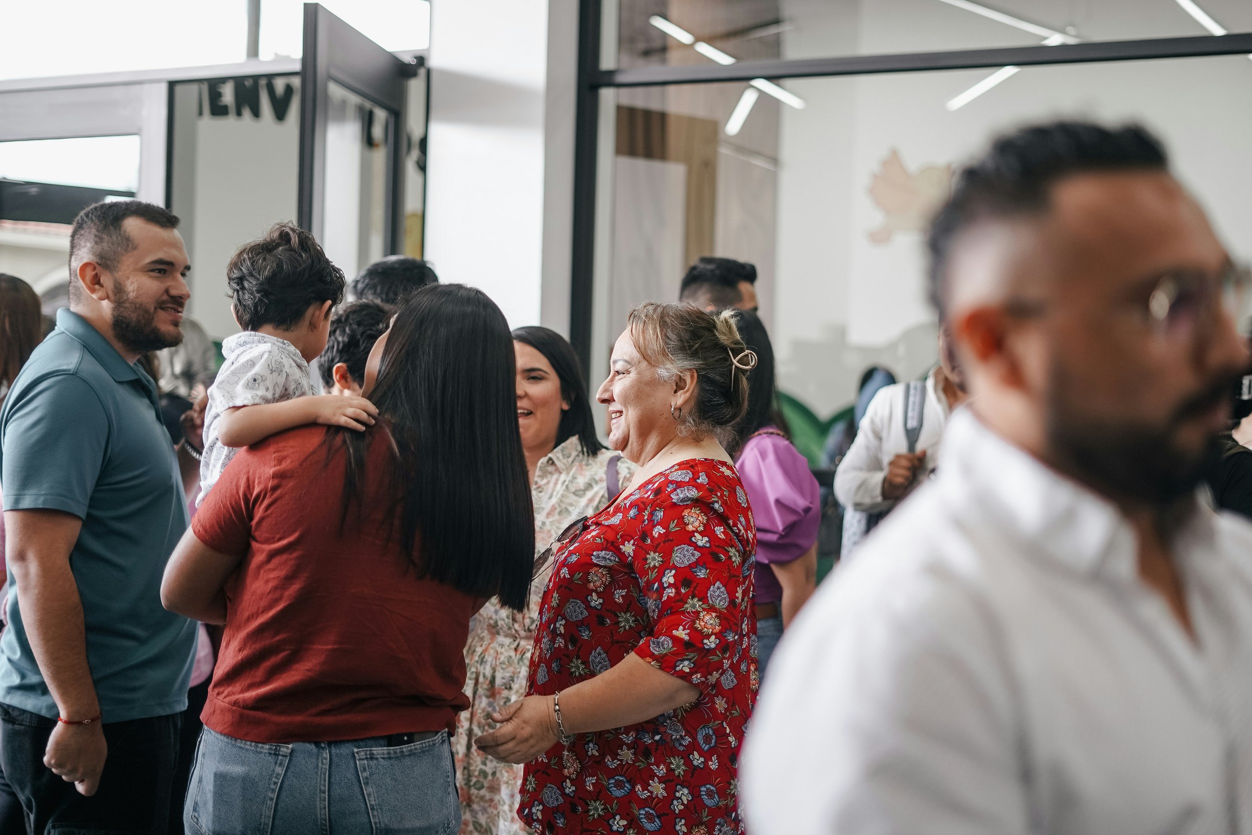 A group of diverse people, including children and adults, gathered indoors, engaging in conversation and smiling, suggesting a social or family event.