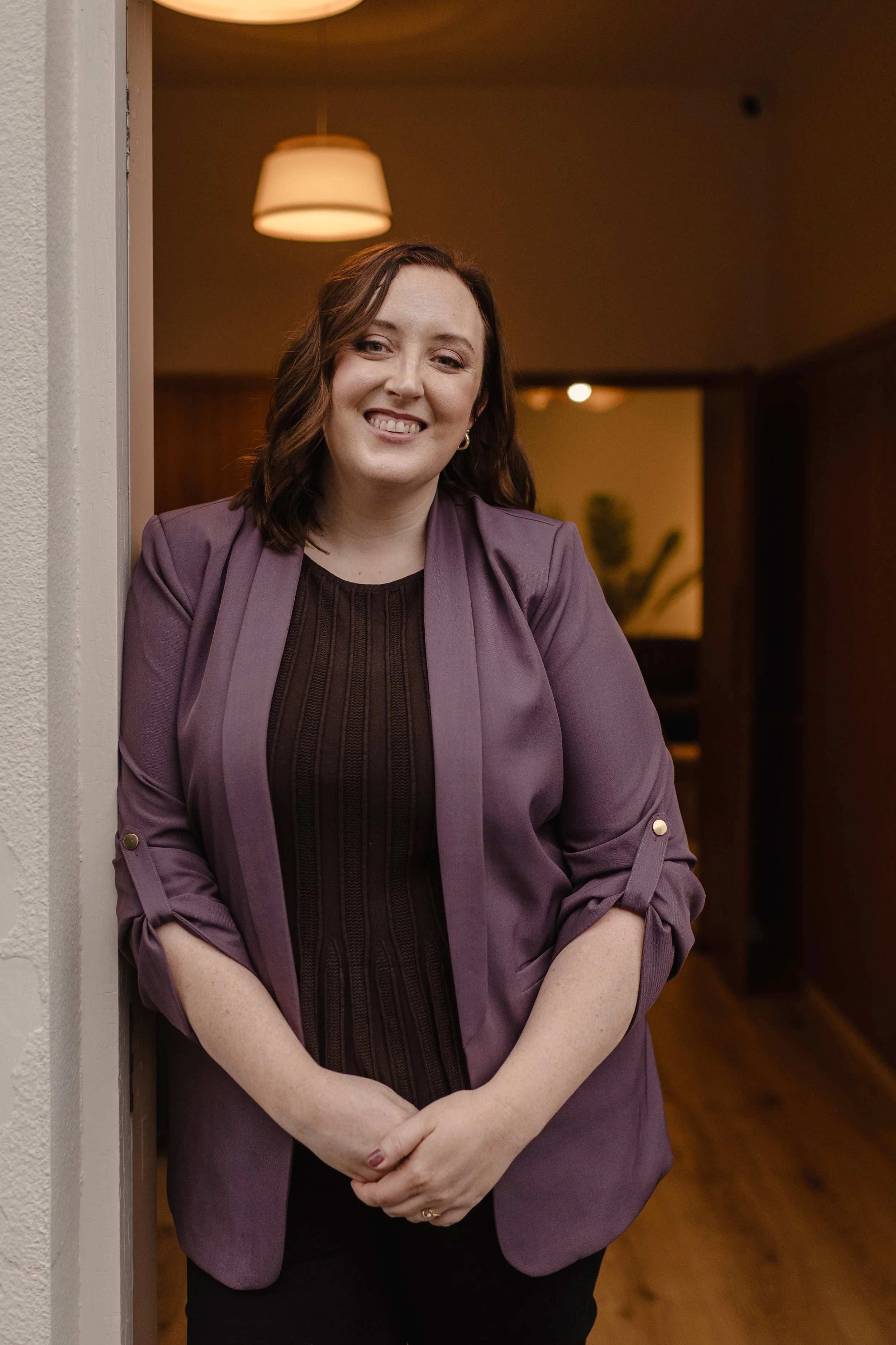 A woman with shoulder-length brown hair wearing a purple blazer and black top, standing indoors near a doorway, smiling with arms crossed in front of her.