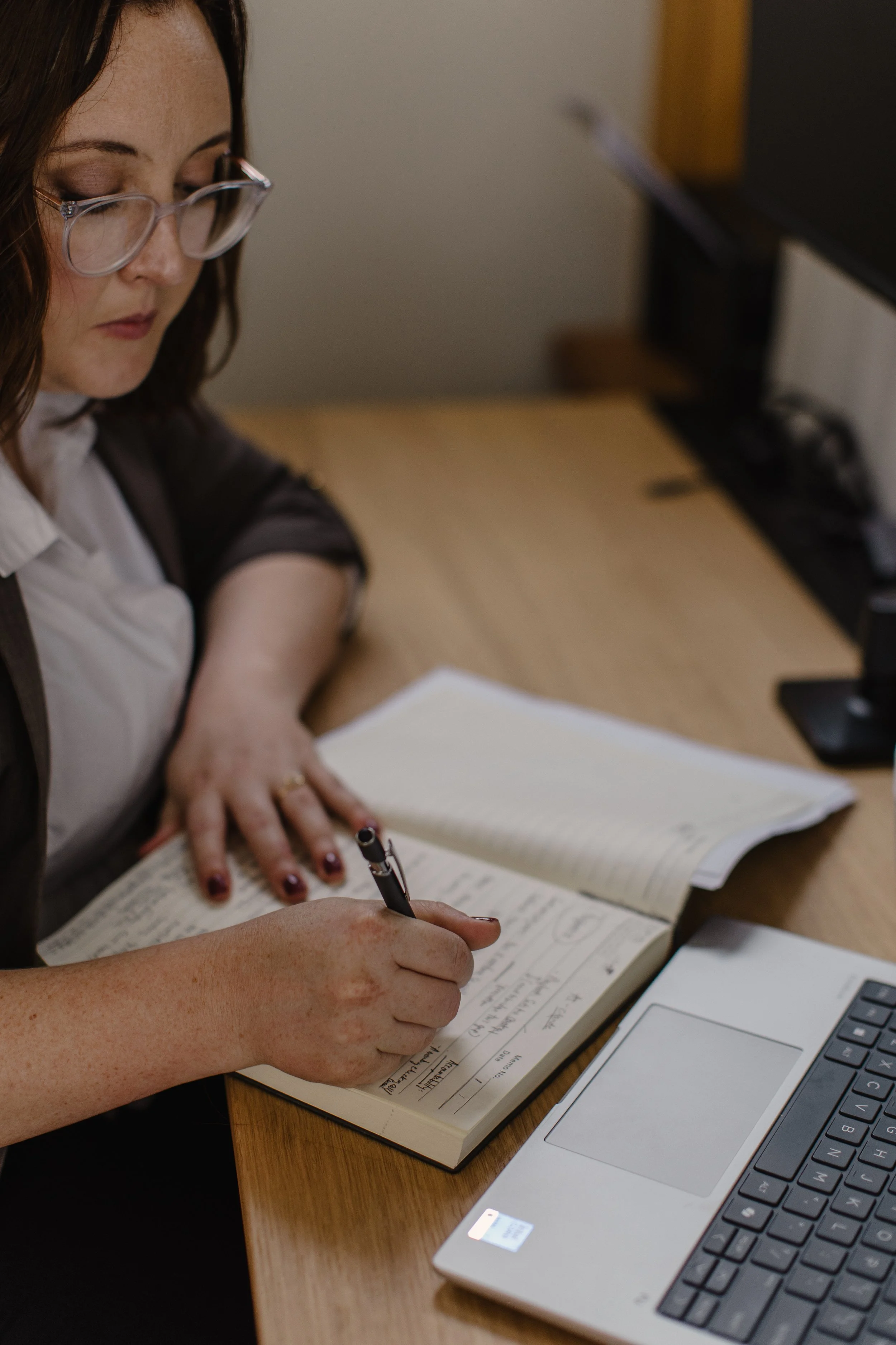 Person taking notes in a notebook at a desk with a laptop and a computer monitor.