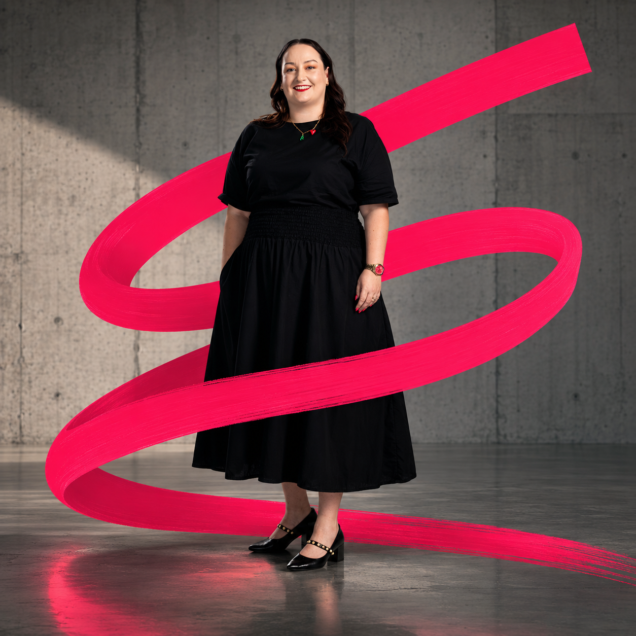A woman wearing a black skirt and top, standing on a concrete floor against a gray wall. She is smiling and has long dark hair. There is a digitally added pink ribbon swirling around her, creating a dynamic visual effect.