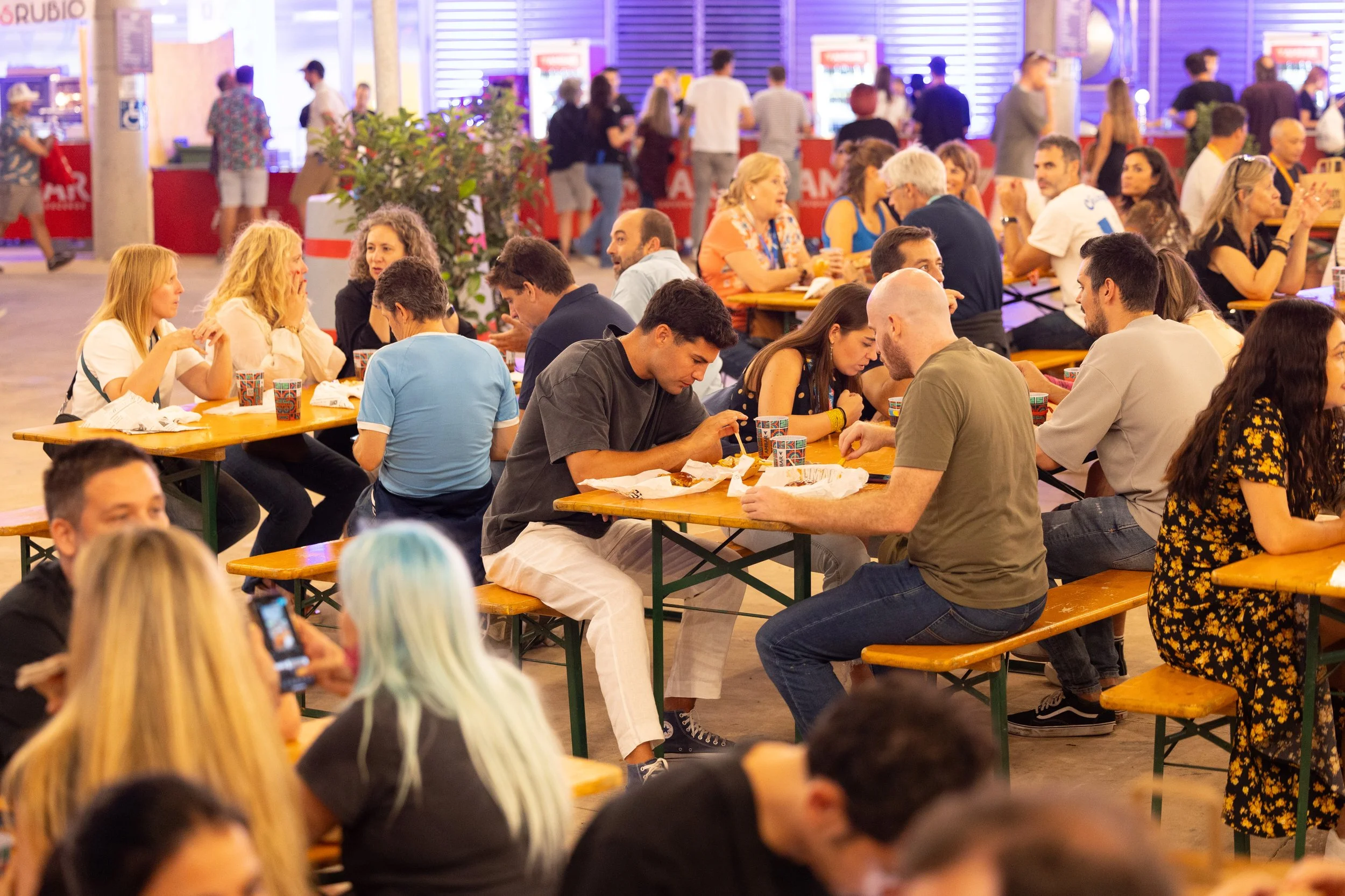 Personas sentadas en mesas de madera comiendo y conversando en un evento social, fondo con una fila de máquinas expendedoras y gente en línea