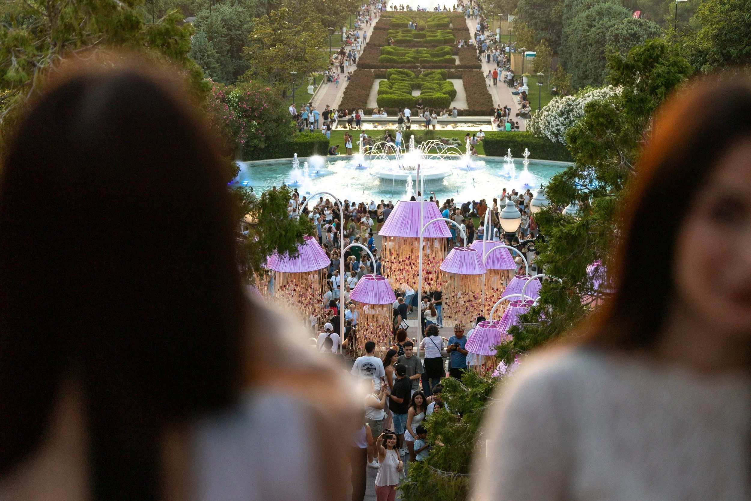 Vista desde un mirador en un parque lleno de muchas personas, con un estanque con fuentes en primer plano y un jardín de diseño en el fondo, rodeado de árboles y vegetación.