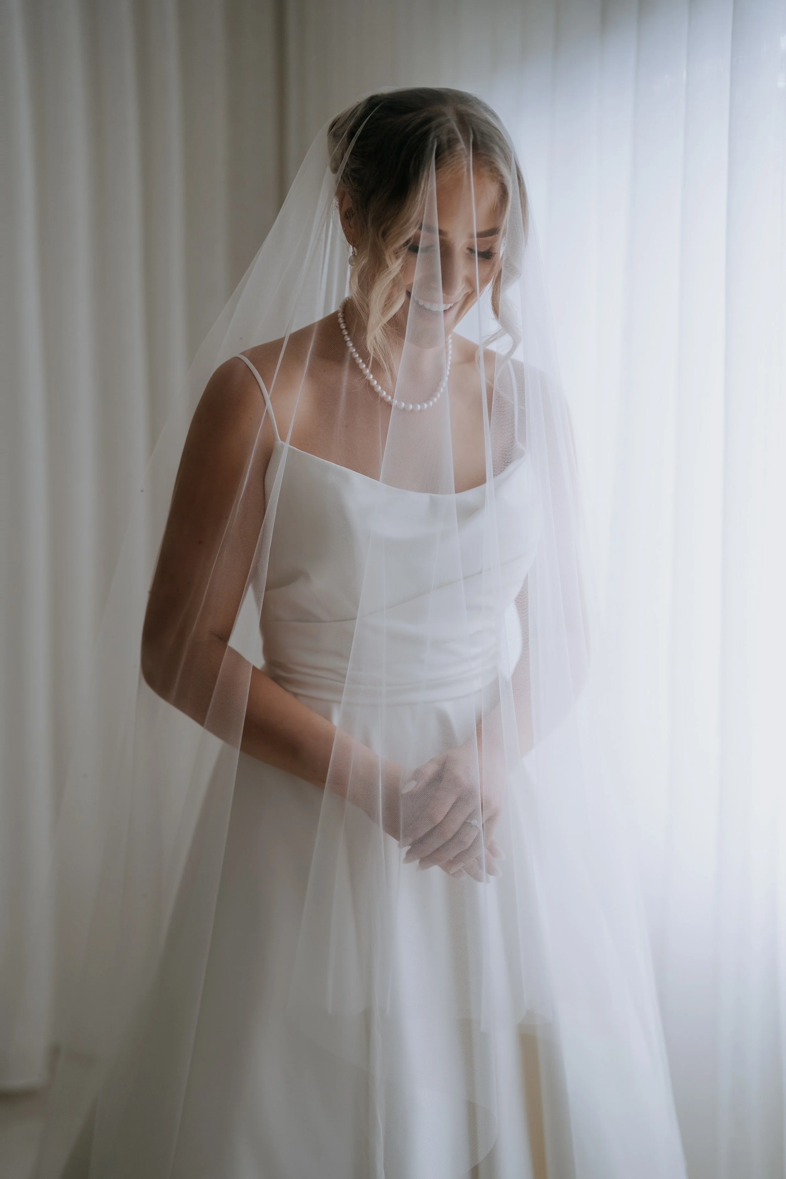 A bride in a white wedding dress with spaghetti straps, wearing a pearl necklace, and a veil, standing behind sheer white curtains, smiling with eyes closed.