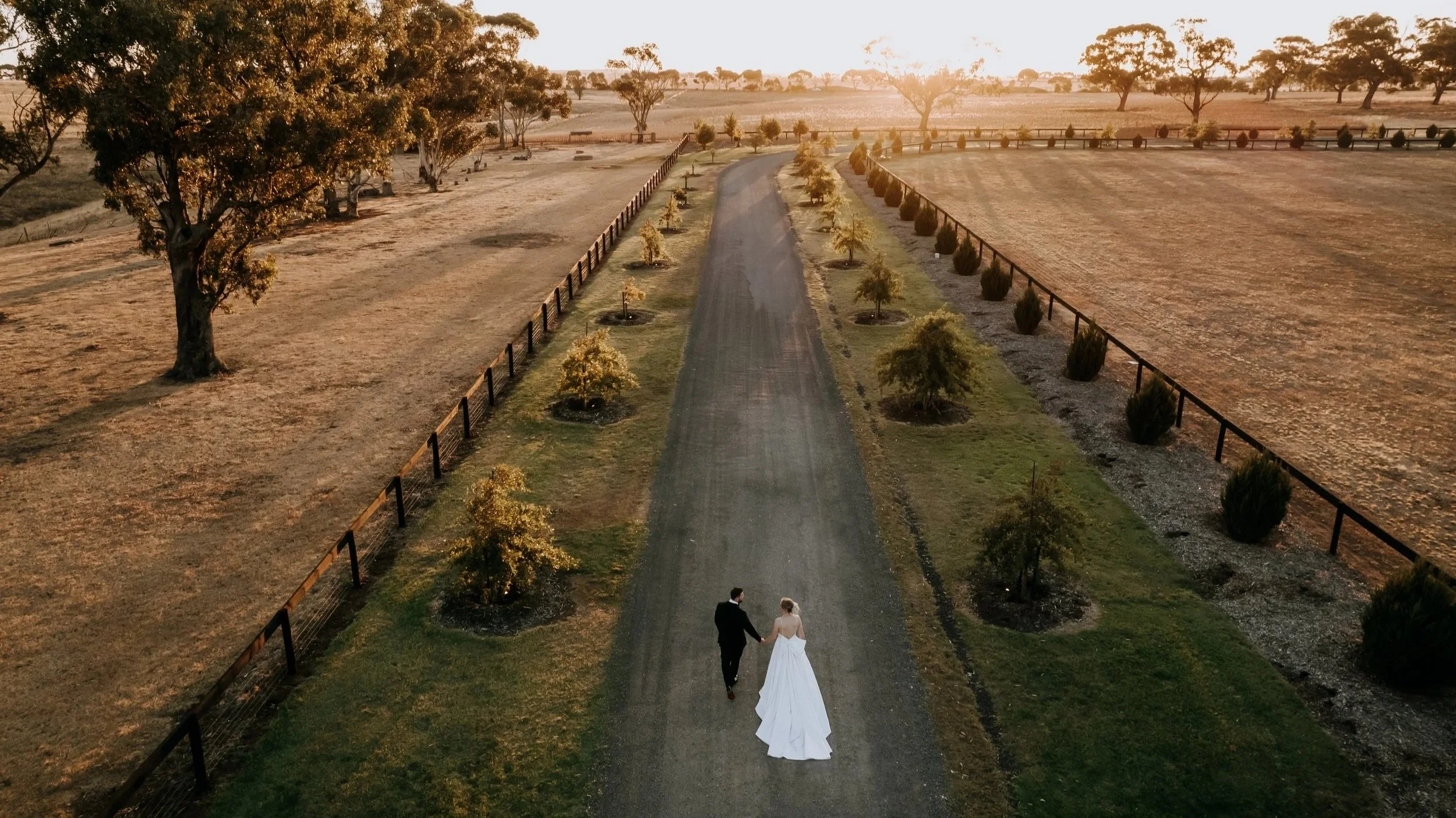 A man and woman dressed in wedding attire holding hands and walking down a dirt path on a farm, with trees and fields at sunset in the background.