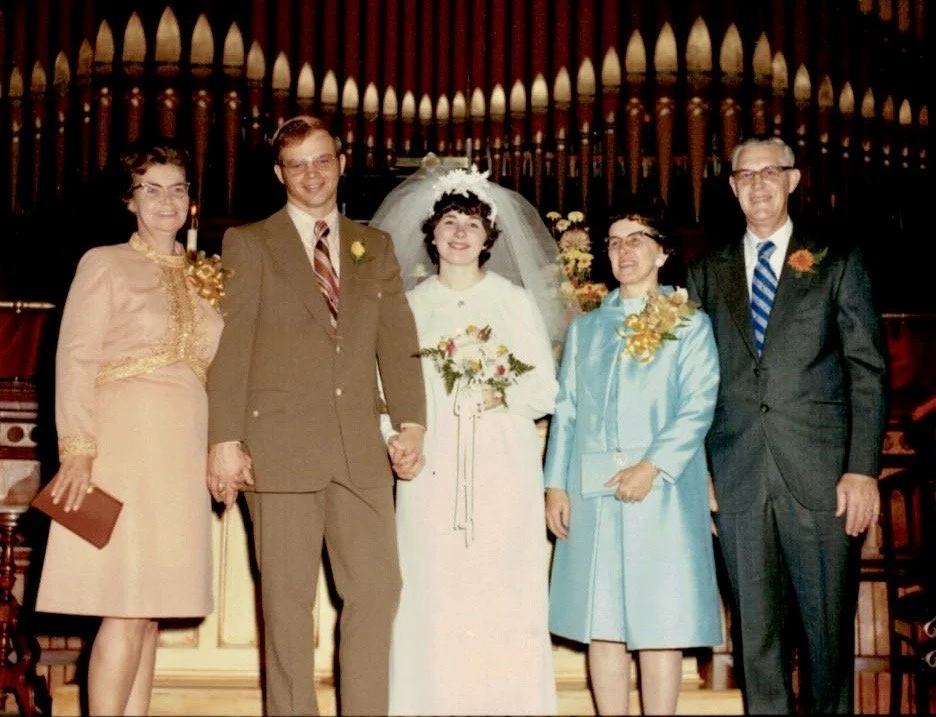 A wedding ceremony with the bride and groom holding hands, surrounded by three family members, indoors with a decorative wooden backdrop.