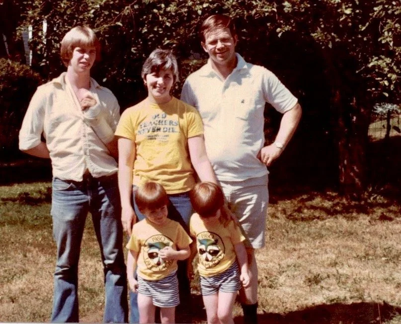 Five children, two boys and three girls, standing outdoors on grass with trees in the background. The children are of varying ages, with the two youngest boys in front wearing yellow T-shirts with a graphic and striped shorts, and the older girls and boy in back dressed in casual clothing, smiling on a sunny day.