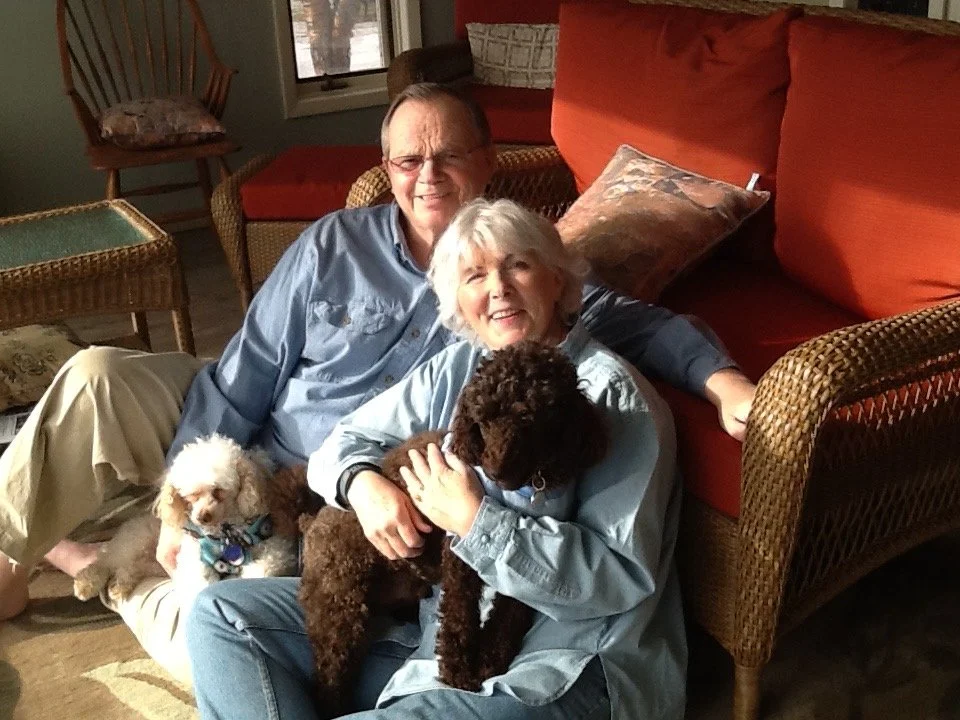 An elderly couple sitting on the floor in a living room, holding two dogs, one white and one brown. The man is wearing glasses and a blue shirt, and the woman has gray hair and is wearing a light-colored shirt. The room has a rattan chair with a cushion and a red sofa behind them.