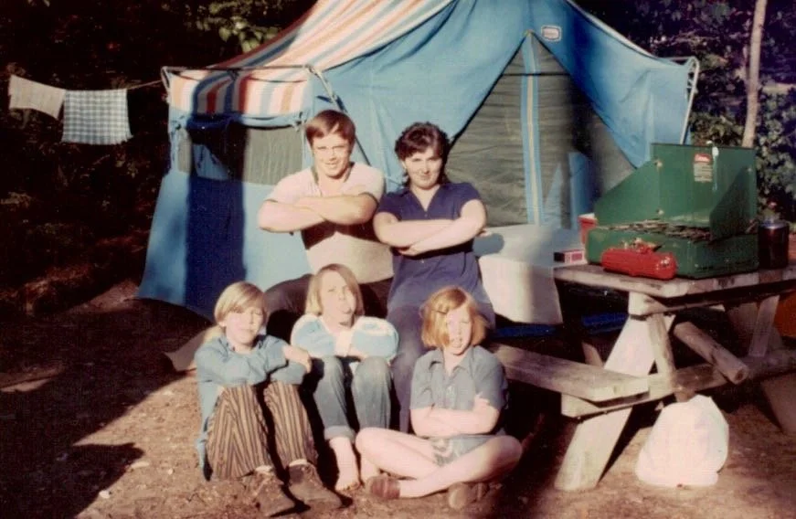 Five children posing in front of a blue camping tent outdoors during daytime, with a picnic table nearby. Two older kids stand with crossed arms behind three younger kids sitting on the ground.
