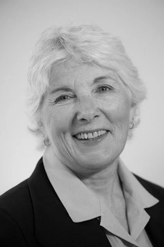 Portrait of an older woman with short, wavy gray hair smiling, dressed in a collared shirt and blazer.