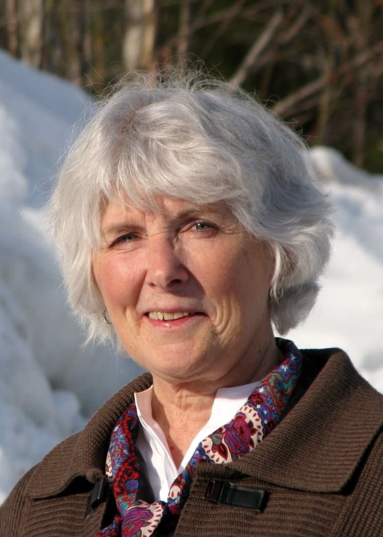 Close-up of an elderly woman with gray hair, smiling outdoors in front of snow-covered landscape and trees, wearing a brown jacket and a colorful scarf.