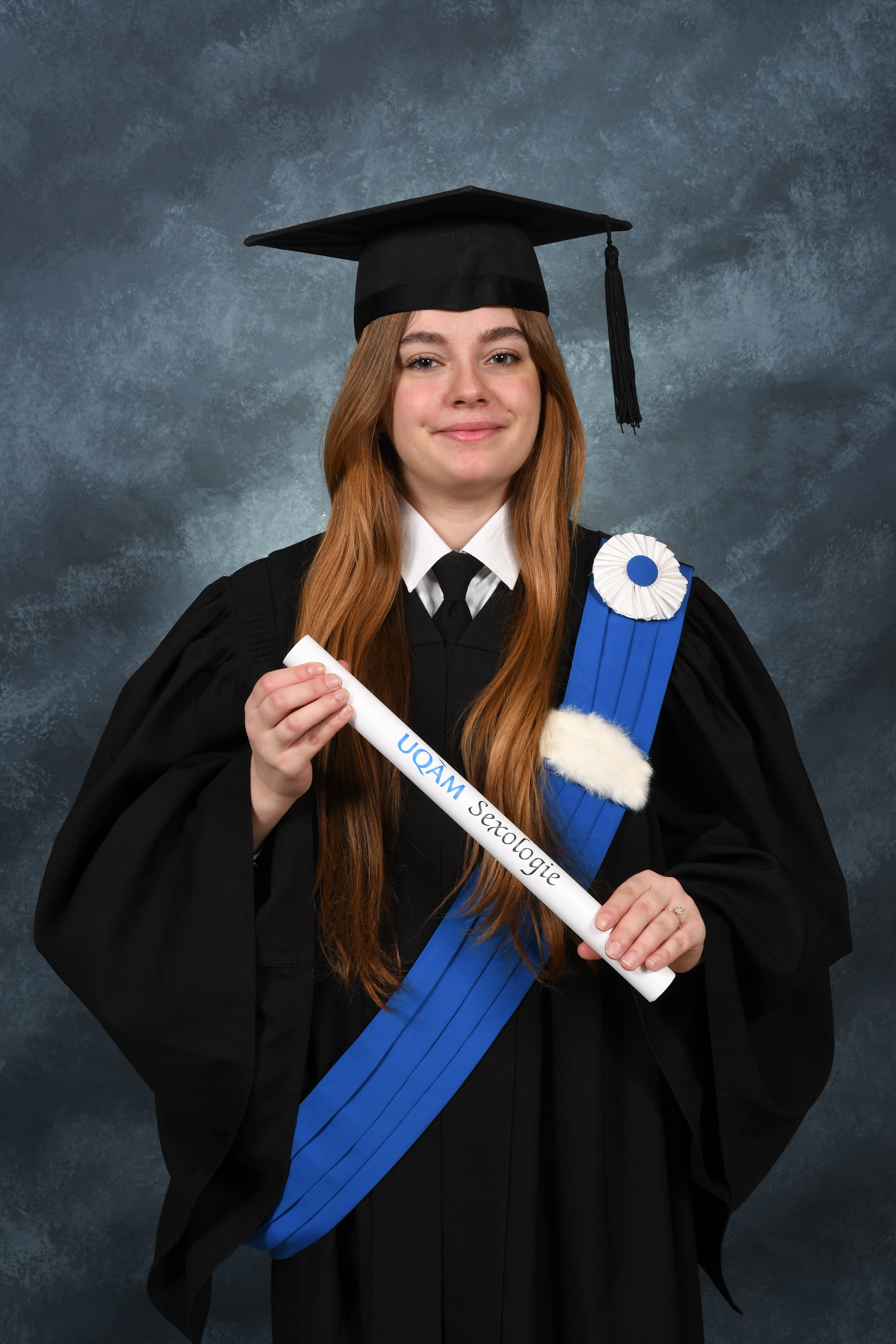 Une jeune femme en robe de graduation noire, portant un bonnet de graduation noir, tient un diplôme blanc et affiche un sourire, avec une rosette blanche à son épaule. Elle porte une bandoulière bleue sur l'épaule.