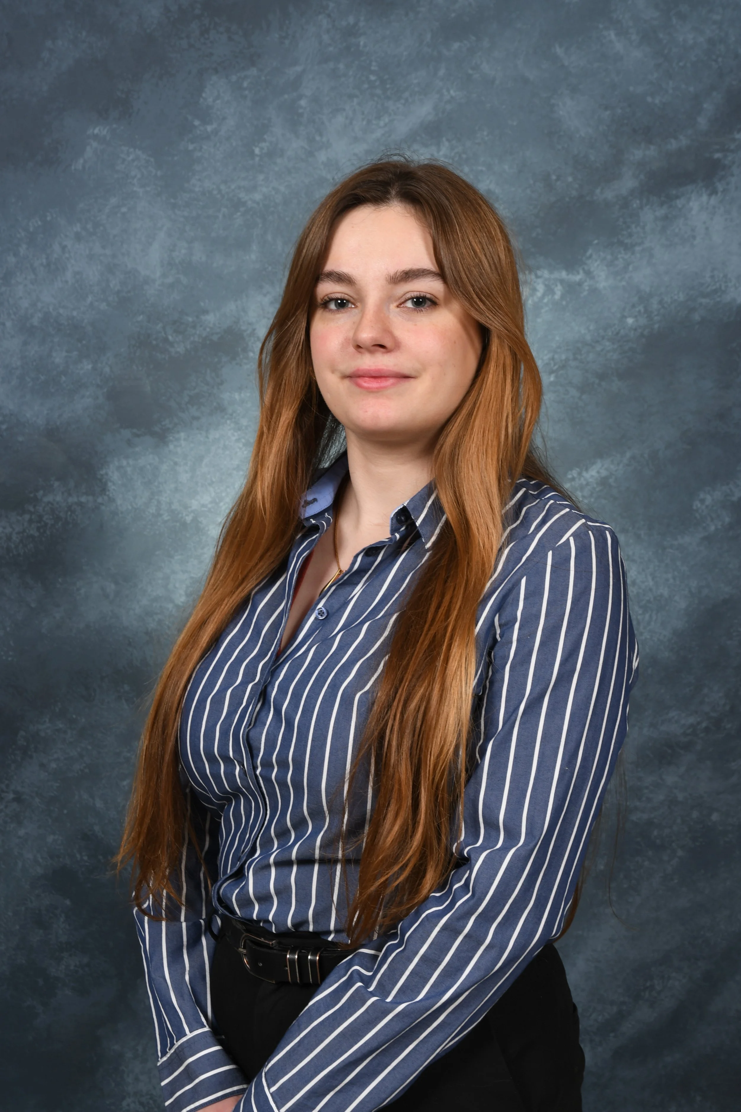 Une jeune femme aux cheveux longs bruns, portant une chemise à rayures bleues et blanches, pose contre un fond gris texturé.