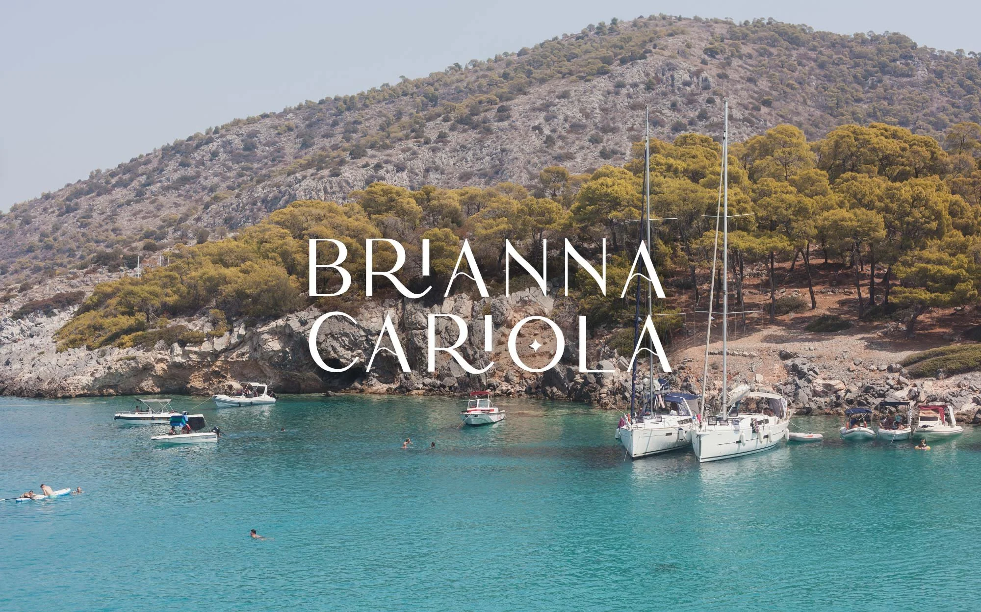 Brianna Cariola photography brand identity stacked logotype overlaid on a travel photograph of sailboats in a turquoise Mediterranean cove with a rocky pine-covered hillside.