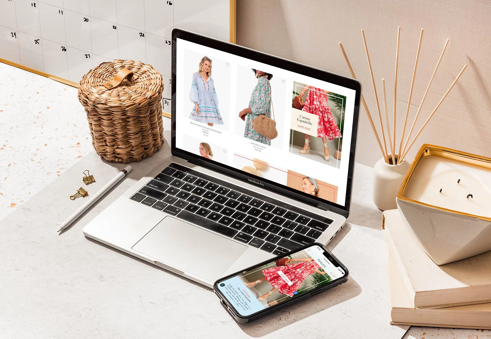 A workspace with a laptop displaying an online fashion store, a smartphone showing the same webpage, a white pen, gold binder clips, a woven lid container, a large calendar, a white pot with reed diffusers, a gold tray, and a beige storage box, all arranged on a light-colored surface.