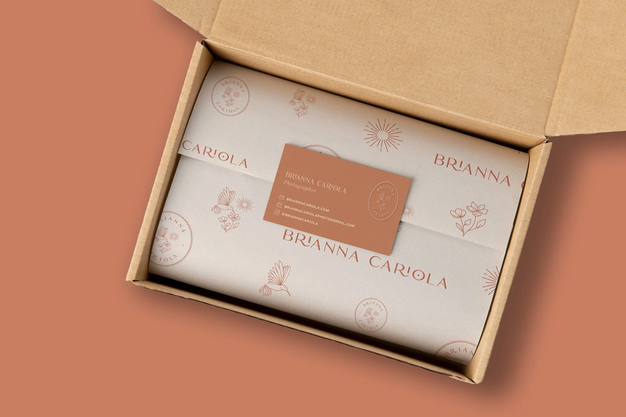 Brianna Cariola photography brand identity business card and branded tissue paper packaging mockup inside an open kraft mailer box, featuring a terracotta business card with the hummingbird logo mark and cream tissue paper with the brand pattern.