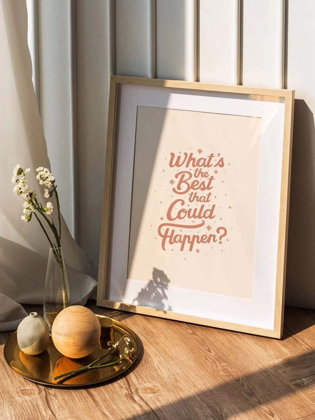 'What's the Best that Could Happen?' retro script typography motivational art print in terracotta pink, displayed in a wood frame leaning against a white paneled wall in warm sunlight beside a glass vase with flowers and a decorative tray.