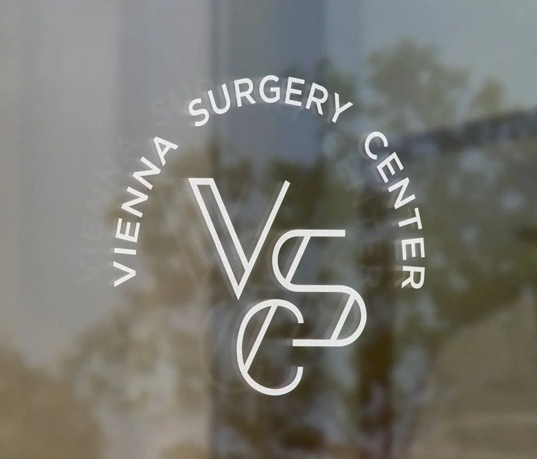 Vienna Surgery Center VSC monogram logo design as a frosted white vinyl decal on a glass door with a warm wood frame, featuring interlocking V, S, and C letterforms in an arched badge layout.