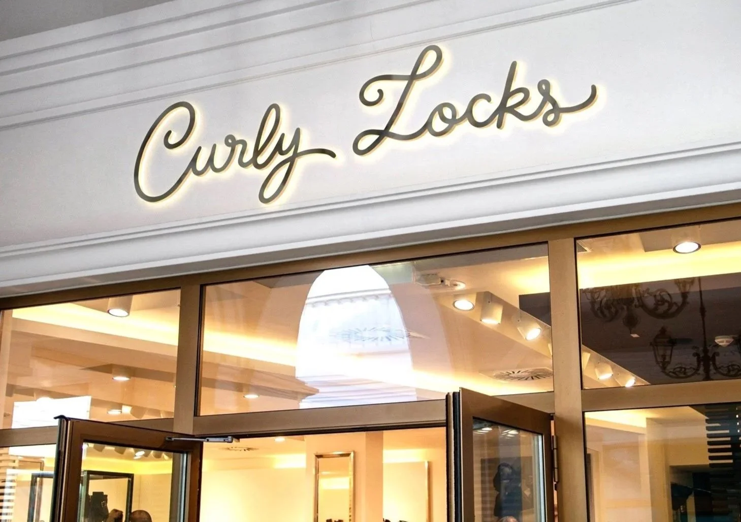 Curly Locks curly hair care brand logo design displayed as an illuminated backlit storefront sign in elegant gold script lettering above glass shop entrance doors.