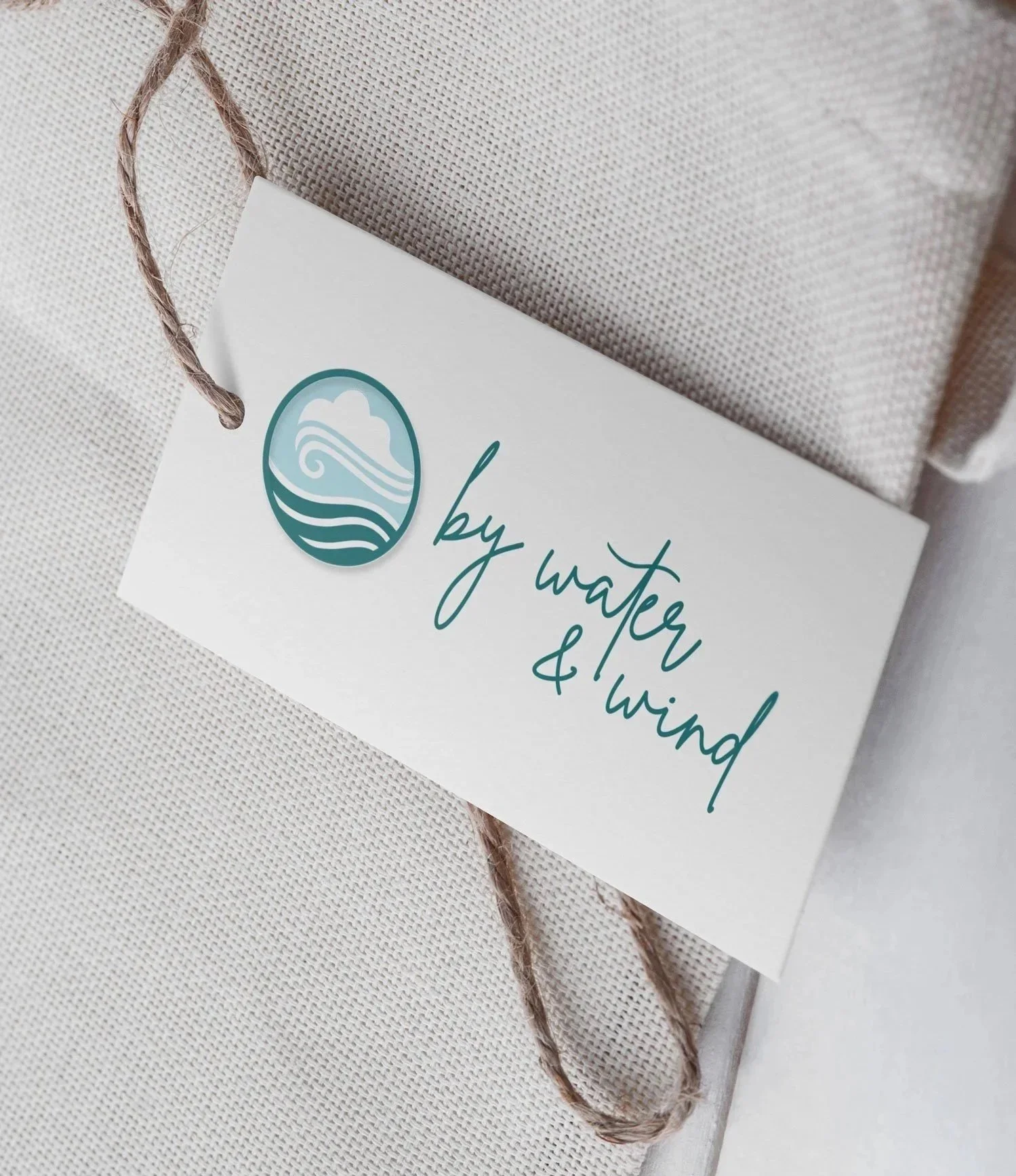 By Water and Wind apparel brand logo design on a hang tag attached to natural linen fabric, featuring a teal oval icon with ocean waves and clouds alongside cursive script lettering.