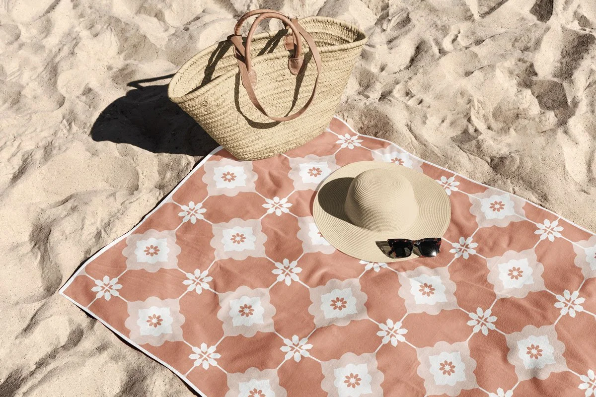 Mediterranean terracotta tile pattern beach towel design mockup laid on sandy beach, styled with a woven straw tote bag, wide-brim sun hat, and sunglasses.