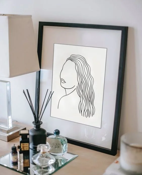Minimal fine line femme woman illustration art print in a black frame, depicting an abstract side profile with flowing wavy hair, displayed on a vanity with perfume bottles and a reed diffuser.