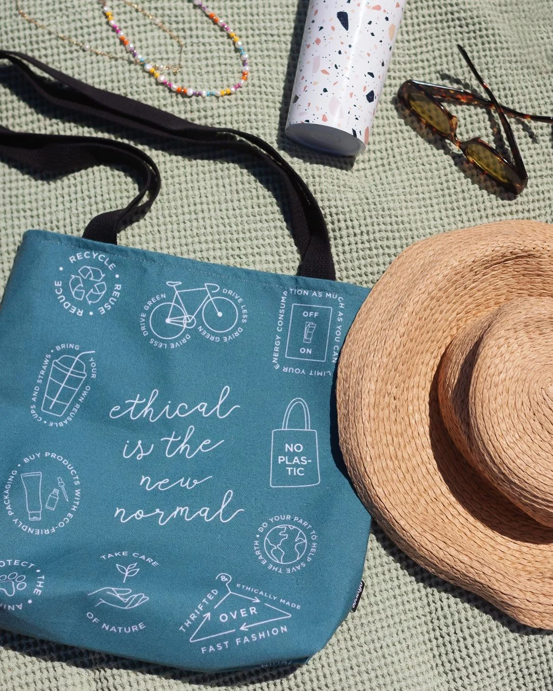 Yasmine Design Co. – sustainability-themed tote bag illustration and graphic design mockup featuring the phrase "Ethical is the New Normal" with eco-conscious icons, styled on a beach blanket with a straw hat and sunglasses.