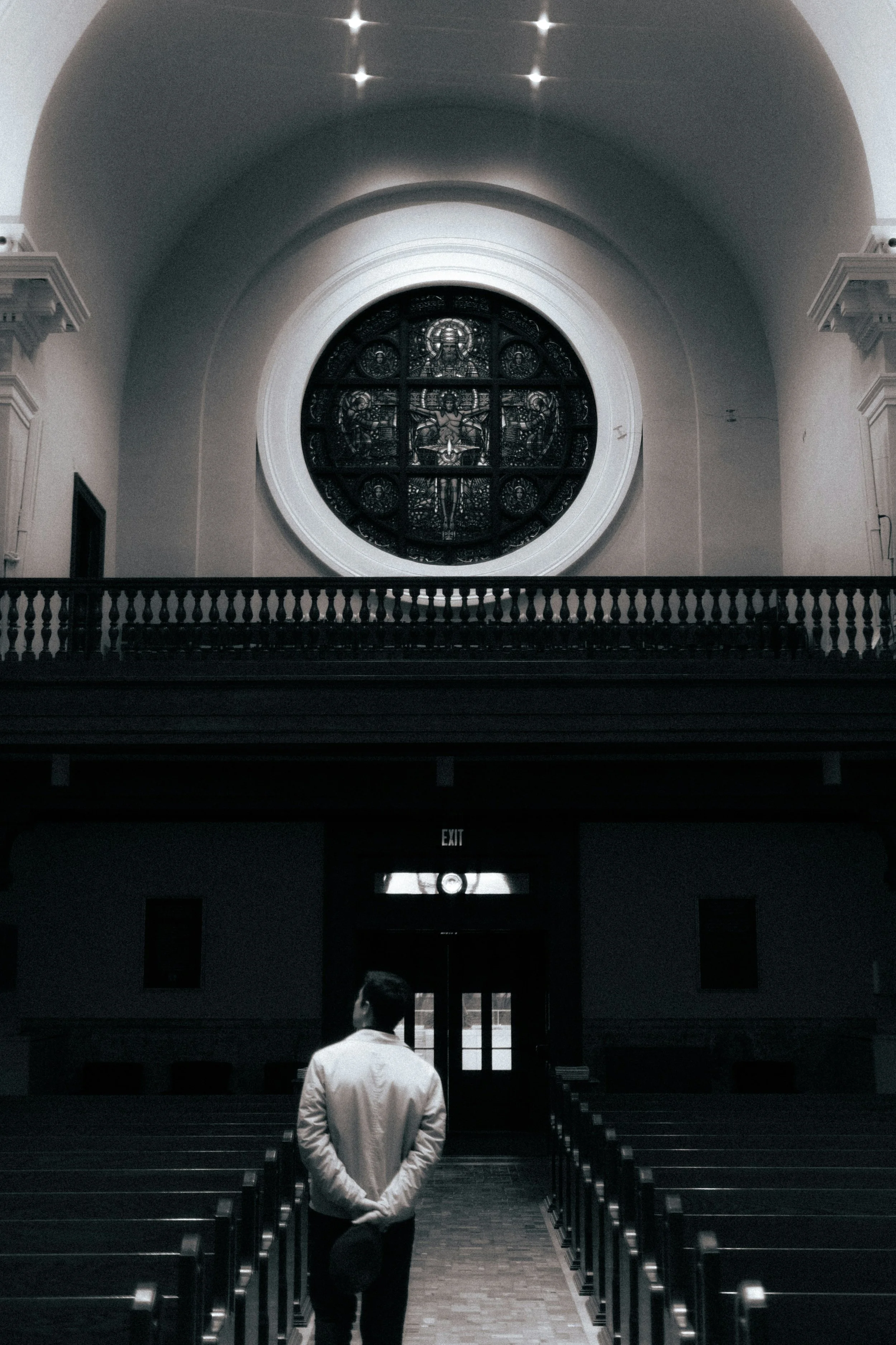 A man in a white jacket and dark pants standing in a church or cathedral, facing away from the camera towards a large circular stained glass window depicting religious figures. The interior has wooden pews and a balcony with a railing, with a sign indicating exit below.