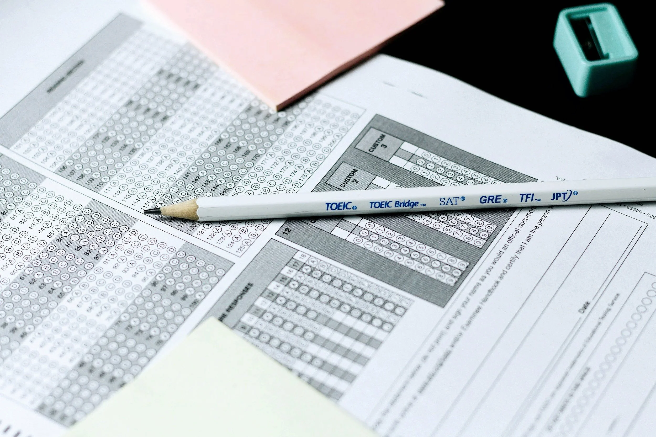 A scanned multiple-choice answer sheet, a mechanical pencil labeled 'TOEIC Bridge,' 'SAT,' 'GRE,' and 'JPT,' a pink note pad, and a teal pencil sharpener on a black desk.