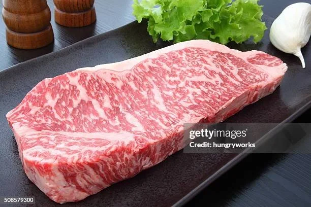 A raw, marbled Wagyu beef steak on a black platter with lettuce, garlic, and salt and pepper shakers nearby.