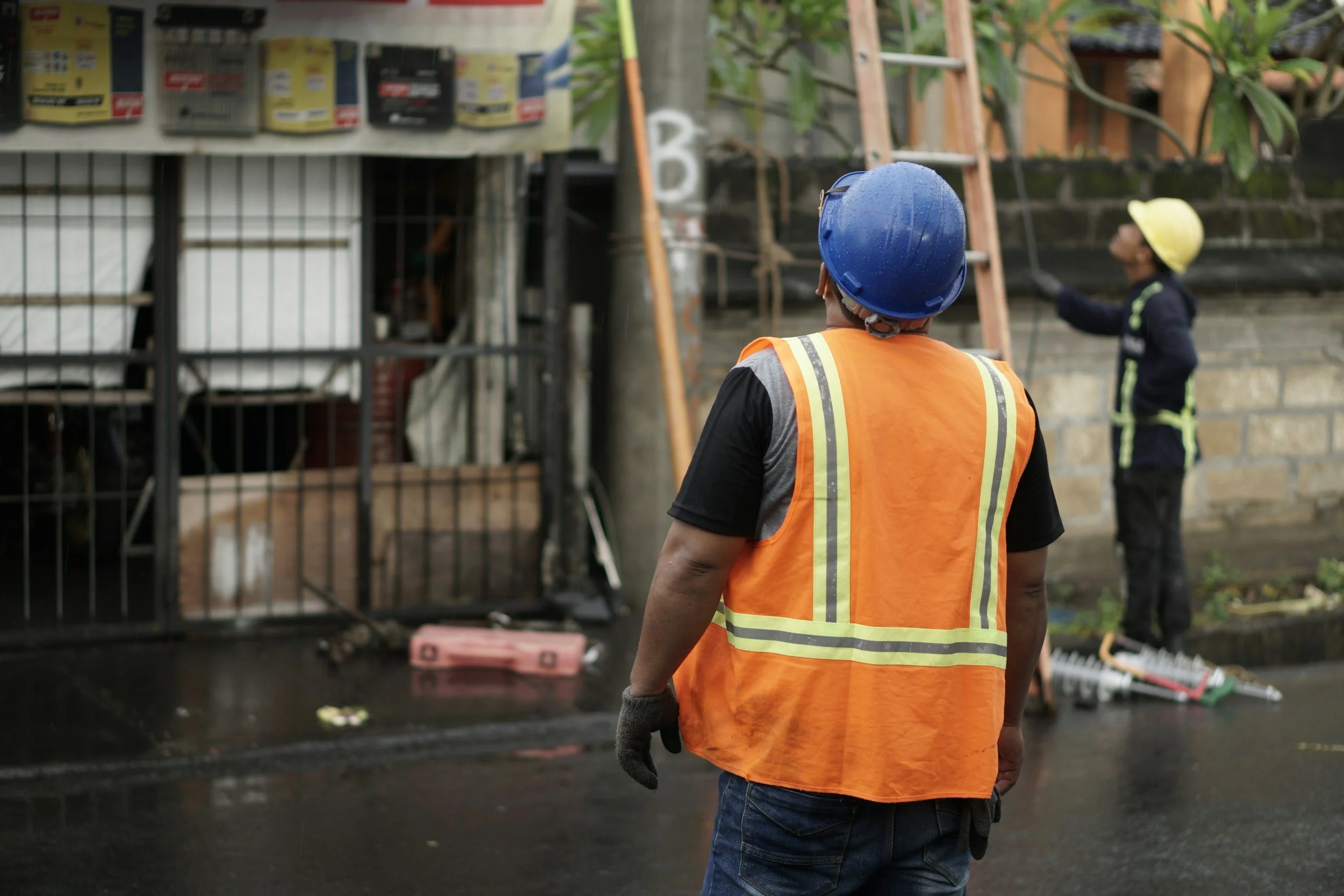 A construction worker wearing an orange safety vest, jeans, gloves, and a blue hard hat looks at a construction site with another worker in a yellow hard hat in the background, along with construction materials and equipment.